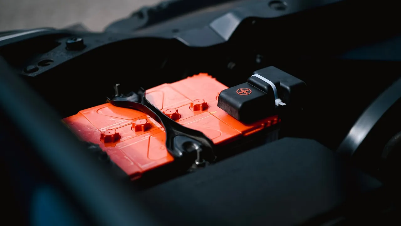 Close-up of a red-top car battery secured by a bracket in an engine bay.